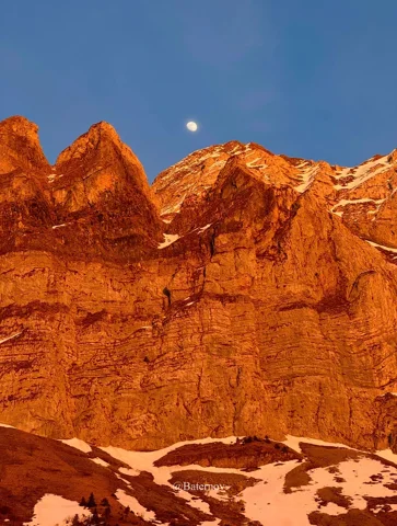 Orange evening light on rugged peaks beneath the moon