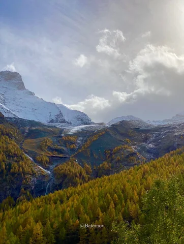 Autumn forest beneath snow-covered mountains and clouds