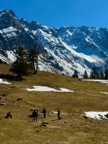 People crossing a mountain meadow below snowy peaks